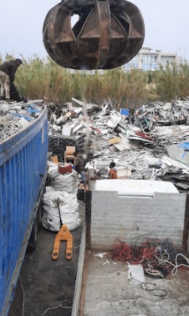 Industrial scrapyard scene with a large metal grapple lowering into a heap of scrap metal beside a blue container and scattered cables.