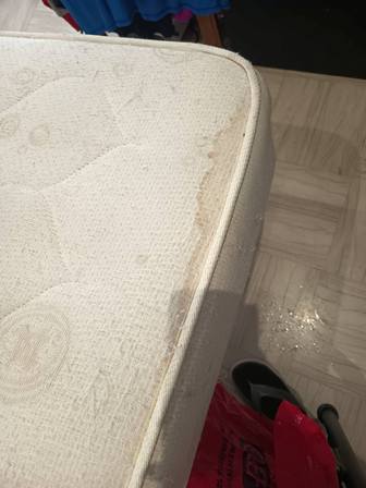 Close-up of a mattress corner with a brown stain along the side, resting on a tiled floor.