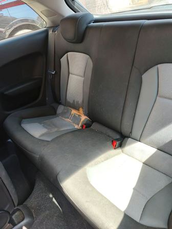 Rear passenger seat in a car with gray fabric upholstery; three-seat bench with two red seat belt buckles visible and sunlight on the right side.