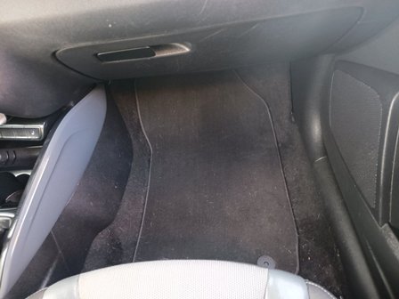 Front passenger footwell in a car showing a black carpet floor mat and part of the center console on the left.