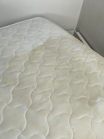 Close-up of a light beige quilted mattress top with a wavy stitched pattern; edge of the bed and a wall corner visible in the background.