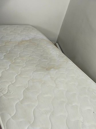 Light-colored quilted mattress with yellowish stains on a bed in a corner room