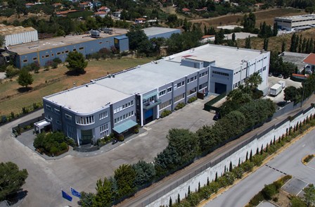 Aerial view of a white-roofed industrial building with multiple connected sections, surrounded by trees and a parking lot.