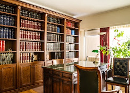 Home office with tall wooden bookcases filled with books, a glass desk, and leather chairs by a sunny window.