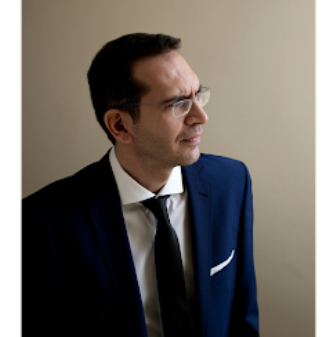 Professional portrait of a man in a navy suit, white shirt, and black tie, wearing glasses and looking toward the right.