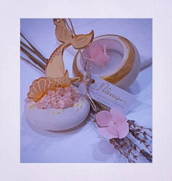 Elegant dessert display featuring a white dome topped with pink crumbles and a gold mermaid-tail chocolate centerpiece, accompanied by pink flowers and a pale tag on a soft purple background