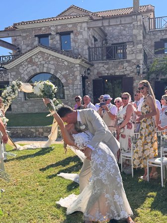 Bride in a lace white gown and a light-colored suit groom kiss outdoors during a wedding, bouquet in hand, with guests watching against a stone villa backdrop.
