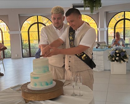 Two men cut a three-tiered cake together at a bright reception hall, one wearing a sash and both in light outfits.
