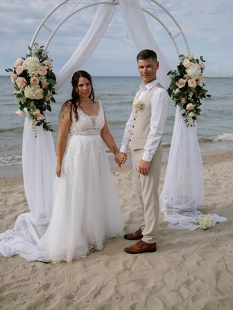 Bride in a white gown and groom in a beige suit hold hands under a flower-adorned arch on a sandy beach with the ocean in the background on their wedding day.