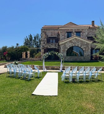 Outdoor wedding setup on a lawn in front of a stone villa, white chairs in rows with a white aisle runner and decorated arch.