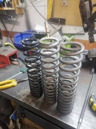 Three large metal coil springs (gray) and two smaller black springs on a metal workbench in a workshop.