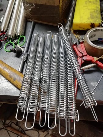 Assorted metal extension springs with hooks laid out on a workbench, with pliers and tape visible in the background.