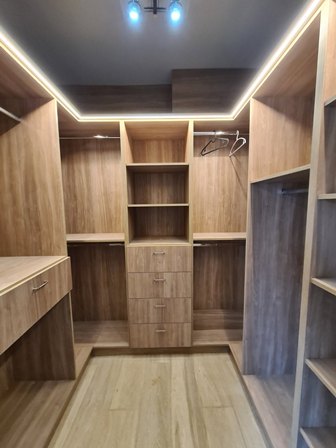 Walk-in wooden closet with central set of drawers, open shelves, and hanging rods on both sides under recessed lighting.