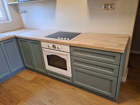 L-shaped kitchen with pale wooden countertop, blue-green lower cabinets, and a white built-in oven with an electric cooktop.