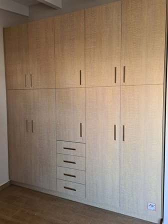 Floor-to-ceiling light wood wardrobe with multiple doors and a central column of drawers in a corner room.