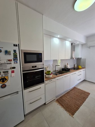White modern kitchen with a tall fridge, built-in microwave and oven, white cabinets, sink, and a beige rug on the floor.