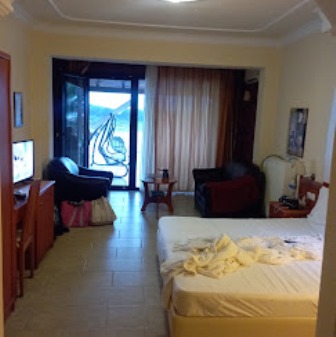 Hotel room with a double bed in the foreground, a seating area, and a balcony door with a sea view.