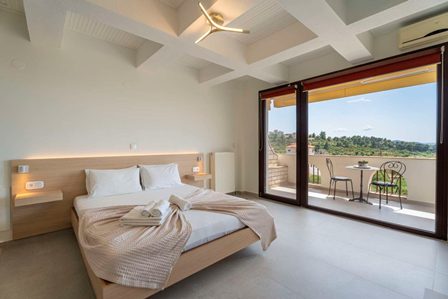 Bright hotel room with a king bed, white linens, and a balcony door opening to a terrace with two chairs and a table overlooking greenery nearby.