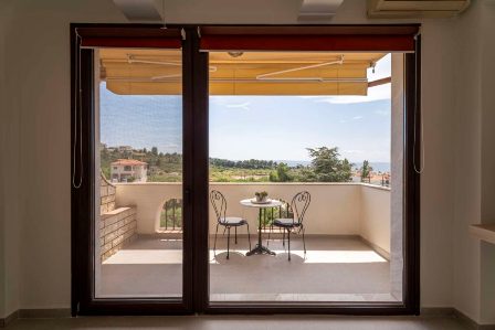Open balcony with a small round table and two chairs under an orange awning, overlooking a green landscape.