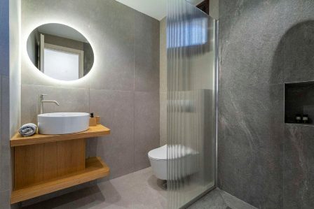 Contemporary bathroom with a round backlit mirror above a wooden vanity with a white vessel sink, glass shower enclosure, and wall-mounted toilet.