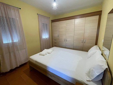 Double bed with white linens and folded towels, facing a large built-in wardrobe; two windows with sheer pink curtains in a yellow room.