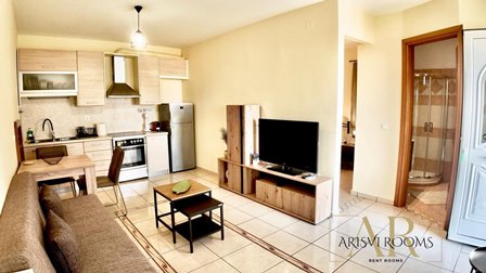 Open-plan living room and kitchen area with a sofa, small coffee table, TV on a wooden stand, and a doorway to a bathroom in the background.
