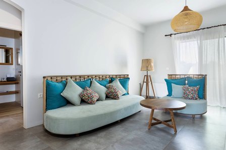 Light-filled living room with a pale blue sofa, matching curved chair, teal and patterned cushions, a round wooden coffee table, and a woven rattan pendant light in the corner.