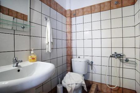 Small tiled bathroom with a white sink, yellow soap dispenser, towel hanging on the wall, toilet with a cloth draped over the seat, handheld bidet sprayer, and a glass shelf.