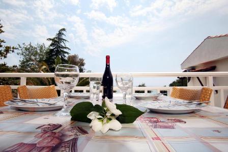Outdoor patio table set for an al fresco meal with a wine bottle, two glasses, and a white flower centerpiece on a patterned tablecloth.