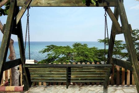 Wooden swing bench on a stone patio overlooking the ocean, with green trees and a clear blue sky.