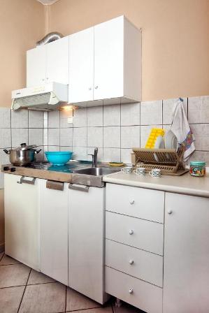 Compact white kitchen with a gas stove and pot on the counter, a blue mixing bowl, sink, dish rack, and white cabinets above and drawers below.