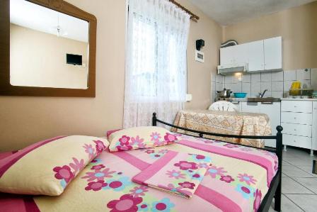 Two beds in a small studio: a pink floral quilted bed in the foreground and a second bed with a yellow cover, with a compact kitchenette in the background.