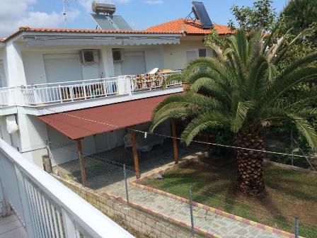 Two-story white house with a red-tiled roof, balcony, and a shaded patio area in a yard with a tall palm tree.