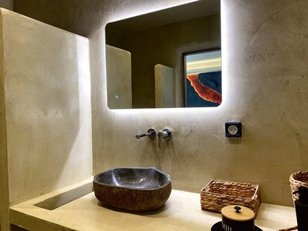Modern bathroom vanity with a dark stone vessel sink, wall-mounted faucet, and a backlit rectangular mirror on concrete walls.