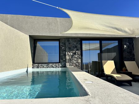 Modern villa with a rectangular pool, glass doors, and two lounge chairs under a white shade sail on a bright day.