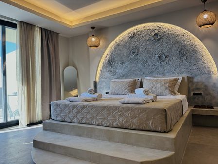 Hotel room with a raised platform bed, round textured headboard, warm lighting, and robes on the bed.