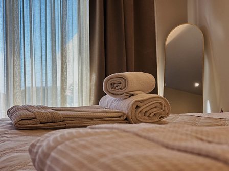 Rolled towels and folded linens on a spa bed by a sunlit window and mirror.