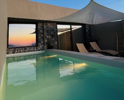 Modern courtyard pool at sunset with two lounge chairs under a shade sail, reflected water, and a stone wall backdrop.