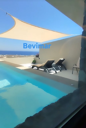 Poolside terrace with two black lounge chairs under a beige shade sail, overlooking the sea on a sunny day.