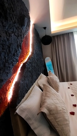 Hotel bedroom with a dark textured wall featuring a glowing lava‑like crack of light; bed has beige pillows, red rose petals, and a snowboard by the window.