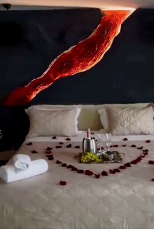 Romantic hotel bed setup with rose petals forming a heart, a towel, and a tray of champagne and fruit, with a red fabric drape overhead.