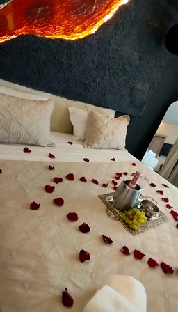 Romantic hotel bedroom: rose petals scattered across a white bed, with a metal tray holding a candle and fruit, and a mirror nearby under a warm amber ceiling light.