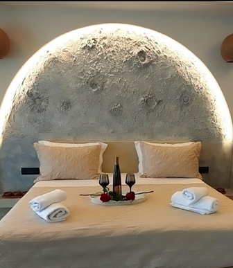 Luxurious hotel bed with white linens and two pillows, a tray of champagne, glasses, roses, and rolled towels against a softly lit curved stone wall.