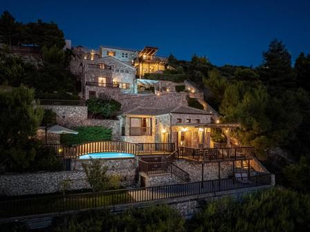 Night view of a multi‑level hillside villa complex with lit terraces and a curved illuminated pool beneath a dark blue sky.