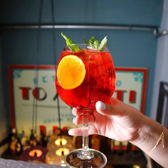 Hand holding a red cocktail with an orange wheel garnish in a stemmed glass, bar setting blurred in the background.