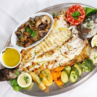 Platter with grilled white fish fillet, roasted potatoes and carrots, zucchini, mushrooms, a lemon wedge, and a tomato slice garnish.