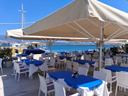Seafront outdoor dining area with white chairs and blue tablecloths under large beige umbrellas, calm blue sea in the background.