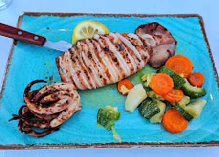 Grilled chicken breast with grill marks on a blue cutting board, served with mixed vegetables (carrots, broccoli, zucchini) and a lemon wedge; knife nearby.