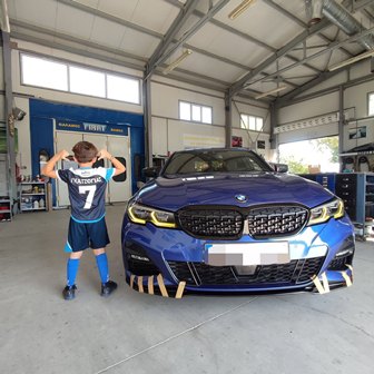 Blue BMW car in a workshop bay, front view with yellow headlights; a boy in a blue soccer jersey with number 7 stands facing the car, hands on head.