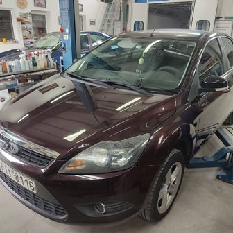 Maroon sedan on a vehicle lift inside an auto repair shop, with tools and shelves visible in the background.